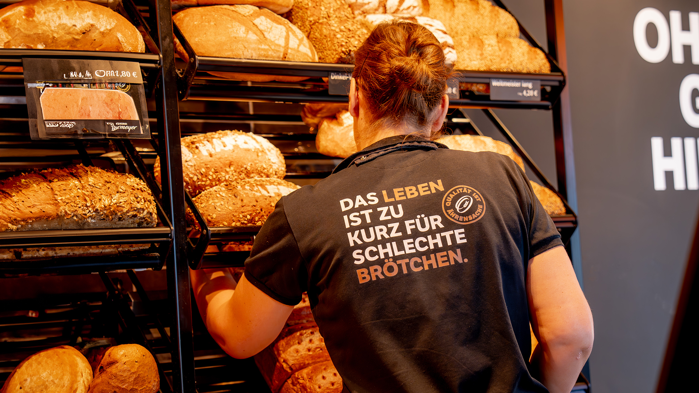 Eine Verkäuferin im Bäckereigeschäft, umgeben von frischem Brot. Auf ihrem T-Shirt steht Das Leben ist zu kurz für schlechte Brötchen Qualität steht im Vordergrund.